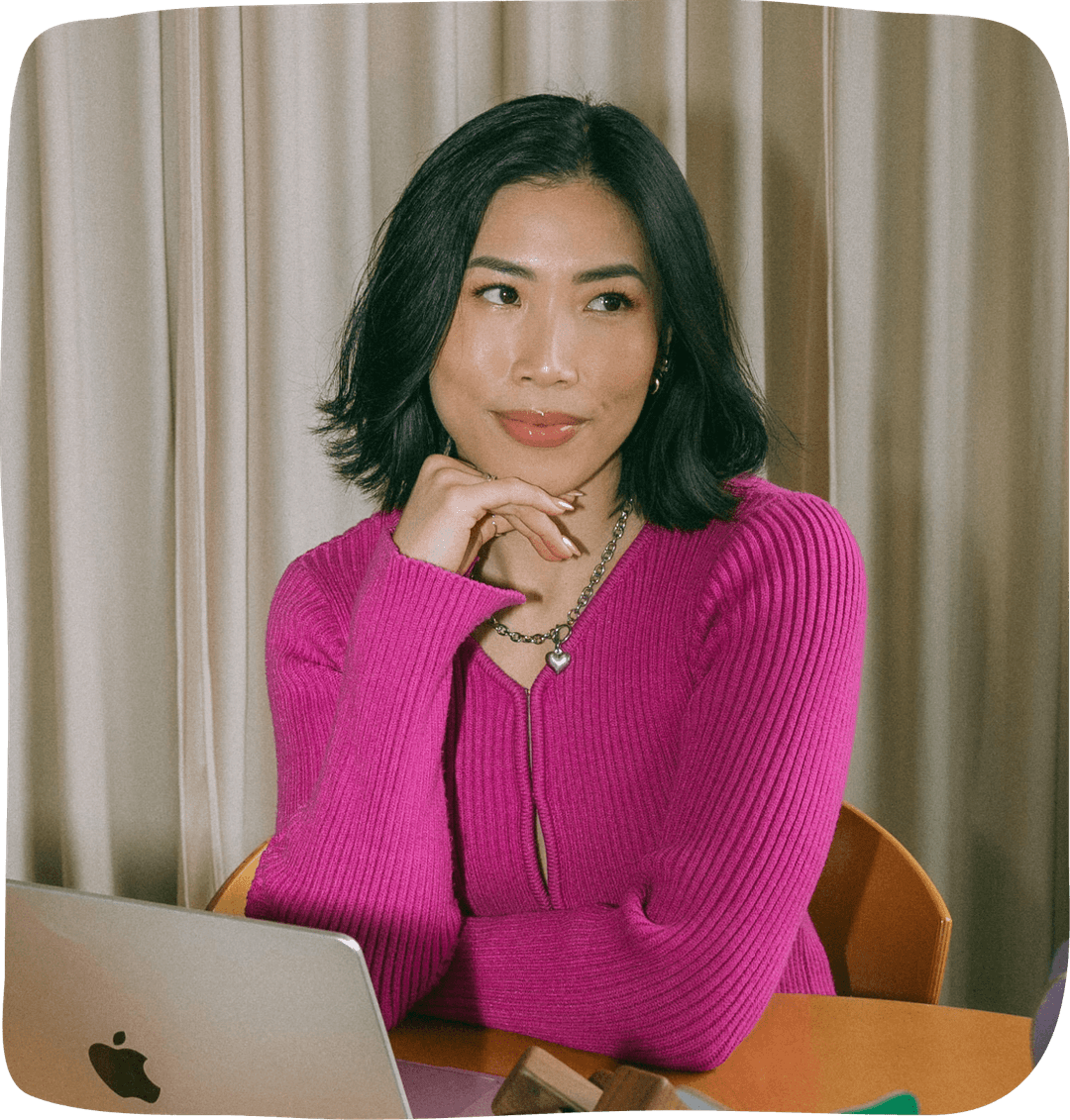 Anna the Cannabinista in a pink sweater resting her elbows on a table with her laptop infront of her, and one hand is placed under her chin as she looks away from the camera. 