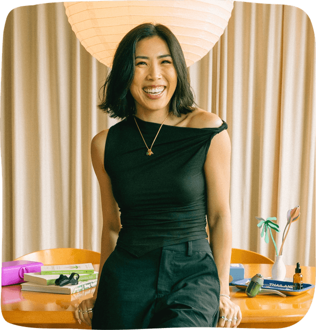 Anna the Cannabinista dressed in a black top and black pants, leaning against a large brown wooden desk with her hands behind her holding herself up as she laughs towards the camera. 