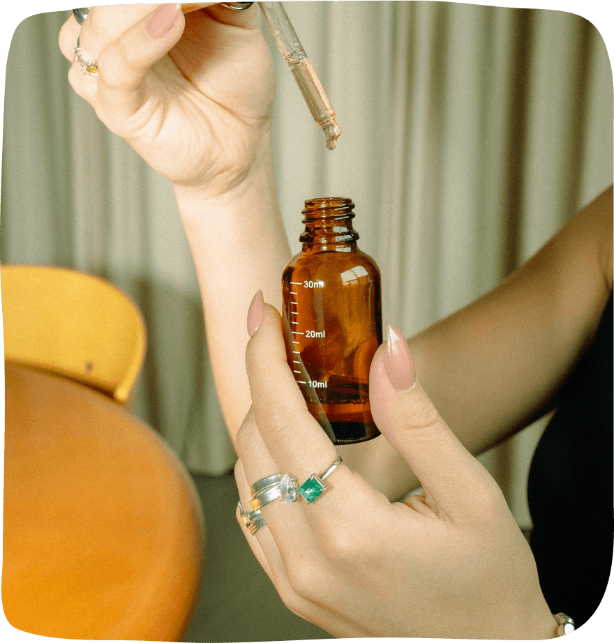 A brown glass cannabis tincture jar is being help up by a hand that has multiple rings on it and the corresponding dropper is being lifted slightly above it allowing drops to fall back into the bottle.